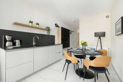 A compact, modern kitchenette with white cabinets and black countertops. A round table with designer chairs provides a dining spot.