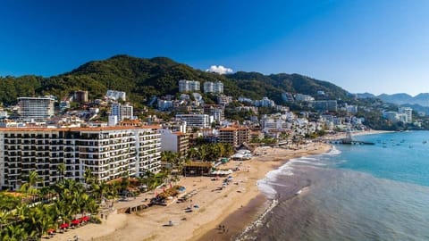 A high-angle aerial view showcasing the beachfront condo complex, sandy shoreline, and surrounding green hillsides.