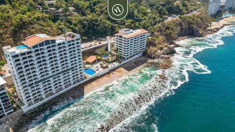 An aerial view of an oceanfront resort featuring multi-story white buildings, a beachfront area with a swimming pool, and waves crashing against the rocky shore.