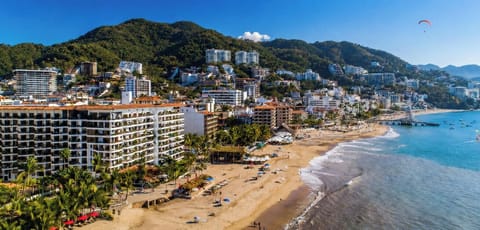 A stunning high-angle shot showcasing the sandy beach, the resort building complex, and the surrounding verdant hills.