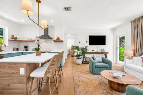 Elegant, sun-filled living room featuring designer coastal décor, plush swivel chairs, a chic white sofa, and custom wood accents, with tropical views and a seamless flow into the gourmet kitchen—perfect for luxurious relaxation and entertaining.