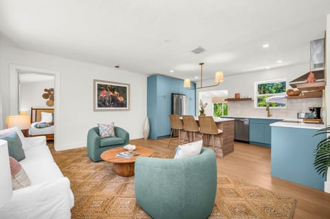 Elegant, sun-filled living room featuring designer coastal décor, plush swivel chairs, a chic white sofa, and custom wood accents, with tropical views and a seamless flow into the gourmet kitchen—perfect for luxurious relaxation and entertaining.