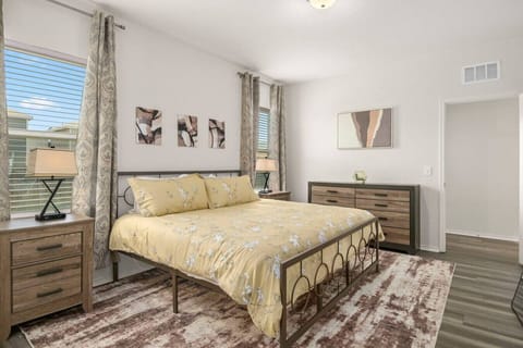 Bright and inviting bedroom featuring a queen bed with yellow floral bedding, matching wood furniture, modern decor, and natural light streaming through double windows with patterned curtains.