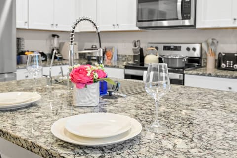 Bright kitchen island with granite countertops and a modern pull-down faucet, set for two with white dishes and wine glasses. A fresh flower centerpiece adds a touch of charm, perfect for enjoying casual meals or morning coffee.