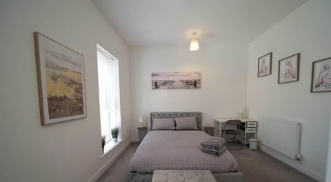 Light and airy bedroom with a cosy double bed dressed in soft grey bedding. Towels are neatly folded on the bed. The room includes a small desk, ottoman, radiator, and framed artworks on the walls, creating a calm and welcoming space.