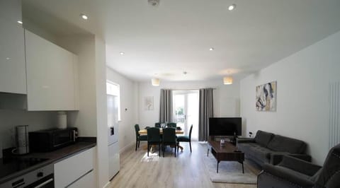 Bright, modern open-plan kitchen, dining, and living area with sleek white units, wooden dining table with green velvet chairs, and comfy grey sofa. Spacious and filled with natural light, ideal for relaxing or entertaining during your stay.