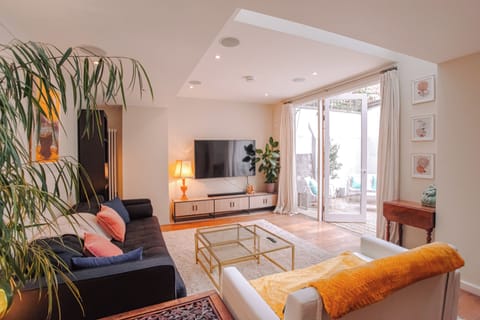 A bright, modern living room features cozy sofas with colorful accents, a glass coffee table, a wall-mounted TV, and large glass doors opening to an outdoor patio.