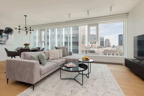 A spacious, modern living room with large windows providing a panoramic view of a high-rise cityscape. The room is furnished with plush gray sofas, a coffee table, and contemporary decor, creating an open and elegant atmosphere.