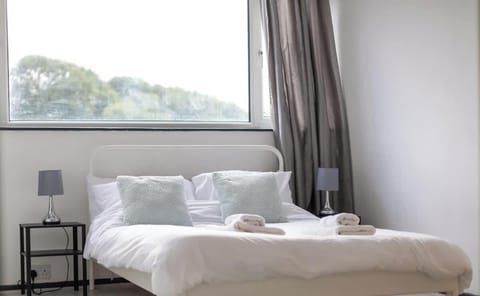 Bright and airy bedroom with a white metal-framed double bed, dressed in crisp white linens and soft cushions. Two bedside tables with matching lamps flank the bed, with a large window providing plenty of natural light and views of greenery outside.