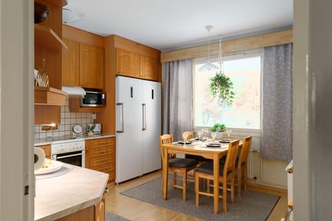 Bright and welcoming kitchen featuring warm wooden cabinetry and modern appliances including a double-door refrigerator, oven, microwave, and toaster. A cozy dining area with a wooden table and four chairs is set by the large window, allowing plenty of na