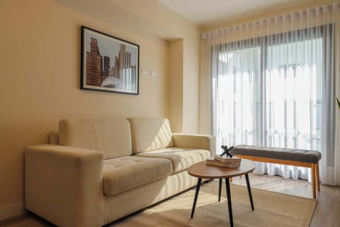 A cozy living room with a beige sofa, coffee table, wall art, and large window with sheer curtains.