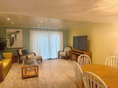 An airy and inviting living space featuring a coastal color palette of seafoam green and soft yellow. The room includes comfortable lounge seating, a large flat-screen TV, and a dining table perfect for group meals, all illuminated by warm, recessed 