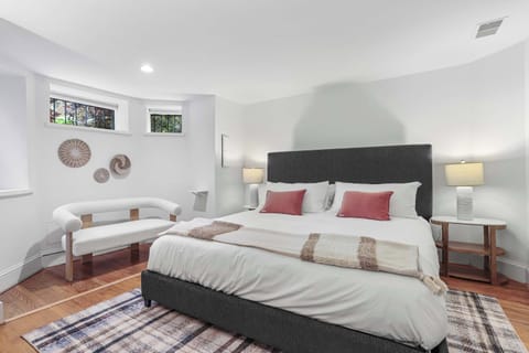 Bedroom with queen sized bed with area rug. The bedroom has nightstands on each side, each with a lamp. There is art and a seat under the windows. 