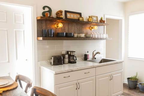 Fully equipped kitchen featuring a refrigerator, sink, stove, toaster, coffee maker, salt and pepper, olive oil, microwave, and essential cooking supplies. A bar table seats two, perfect for shared meals. Handcrafted rustic floating shelves, vintage touch