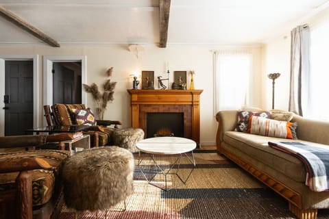 Living room with a cozy country-cabin vibe, featuring a faux fireplace, natural fiber rugs, and a chaise lounge. Handcrafted red oak table, pine accent wall with antique sliding barn doors, built-in reclaimed shelving, and a curated book selection blend m