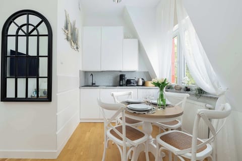 A modern kitchenette with white cabinets and a small dining area. Sheer curtains soften the daylight.