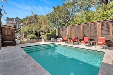 Inviting pool area with stone decking, red cushioned loungers, and privacy shutters, looking back toward the lush garden and charming home for a seamless indoor-outdoor retreat.
