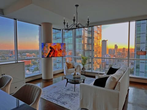 This photo showcases a stunning modern high-rise apartment living room bathed in the warm glow of a sunset. The space features floor-to-ceiling windows on two sides, offering breathtaking panoramic views of a vibrant cityscape.