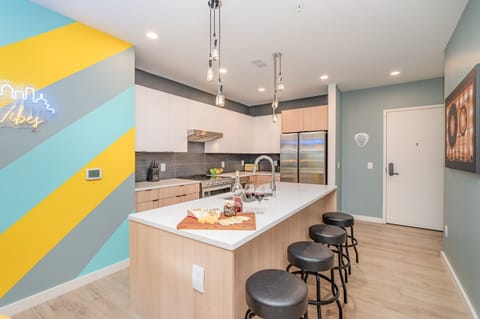 Full kitchen with stainless steel appliances, pristine white countertops, light wood and white cabinetry, and a spacious island with breakfast bar seating for four.