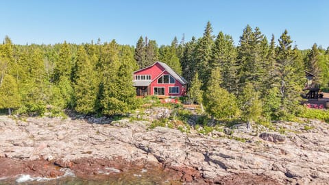 A charming red cabin nestled among evergreens along a rocky shoreline, offering peaceful waterfront seclusion and natural beauty.