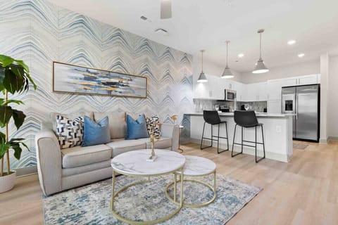 Modern Living Space – Relax in this stylish open-concept living area featuring a chic sofa, marble-top tables, bold accent wall, and seamless flow into a fully equipped kitchen with stainless steel appliances and bar seating.