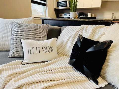 Cozy up on our comfortable sofa, adorned with festive `LET IT SNOW` and plush decorative pillows. A soft white throw blanket invites relaxation. Enjoy a glimpse of our modern kitchen in the background, creating a warm and inviting atmosphere.