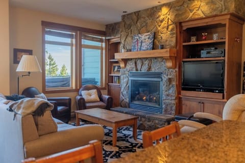 The cozy living room at this Big White chalet features a stone fireplace, leather armchairs, and a beige sofa—perfect for unwinding. Large windows frame snowy views, while a TV and books add relaxation.