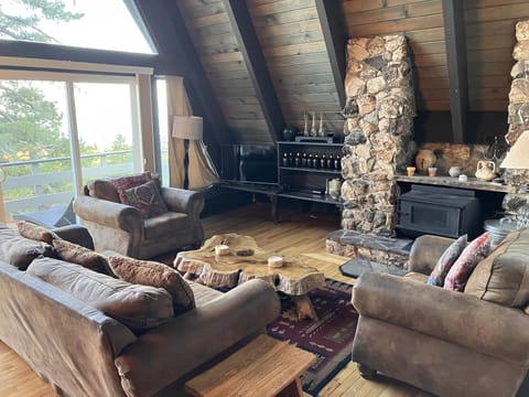 Rustic living room with stone fireplace and forest views through large windows.