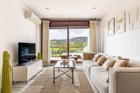 Salón amplio con sofá en L, cojines claros, televisor, chimenea, aire acondicionado, mesa de centro, comedor con seis sillas, lámparas colgantes de mimbre, ventanal con acceso a terraza amueblada y vistas a colinas verdes.
