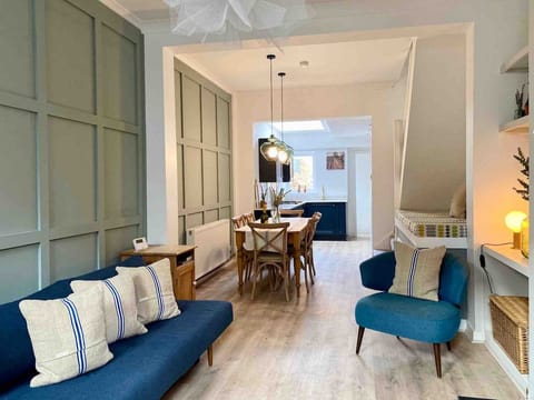 Bright living room with a blue sofa bed and striped pillows, paired with a matching armchair. Muted green panel walls, warm wood floors, and pendant lighting create a stylish, versatile space for lounging or hosting.