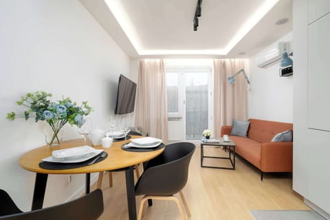 A bright living and dining area with a table set for meals and a sofa in the background. Natural light fills the space, creating a warm and inviting atmosphere.