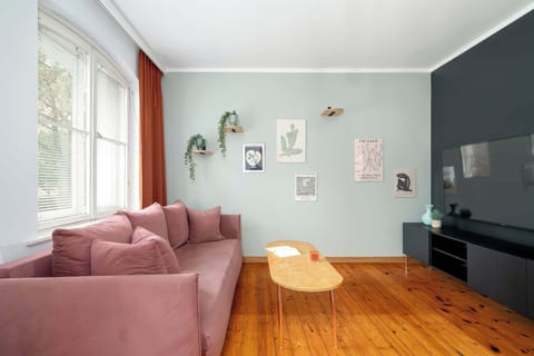 A cozy living room with a muted green accent wall and a comfortable pink sofa. A small round coffee table sits in front, with natural light streaming through the window.