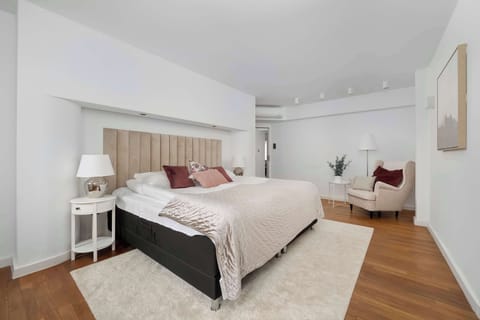 A modern bedroom with a large bed, white walls, and light wood flooring.