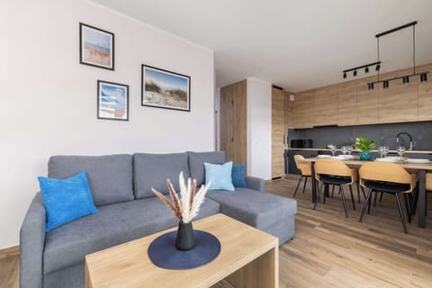 A contemporary living room featuring a gray sofa, a television, and framed artwork on the wall, with a glimpse of a kitchen area in the background.