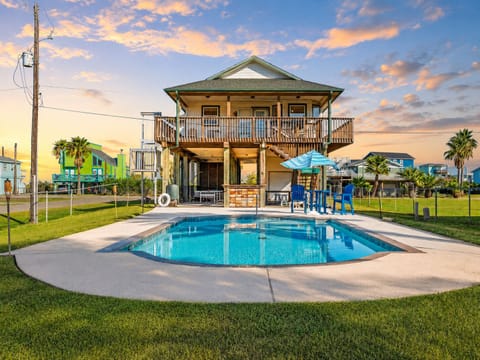 Exterior view of Galveston vacation home with private backyard pool, fenced outdoor space, elevated deck, patio seating, and sunny coastal curb appeal.