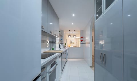 Narrow kitchen with sleek white cabinets and appliances, leading to an open space.