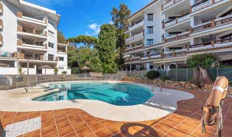 A clear swimming pool surrounded by tiled flooring, with apartment buildings and lush greenery in the background.