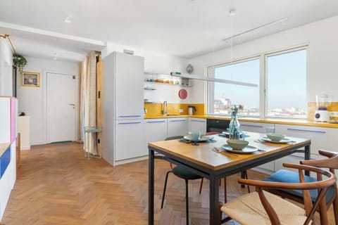 A dining space with a wooden table, four chairs, and large windows. The orange lighting fixtures create a warm tone.