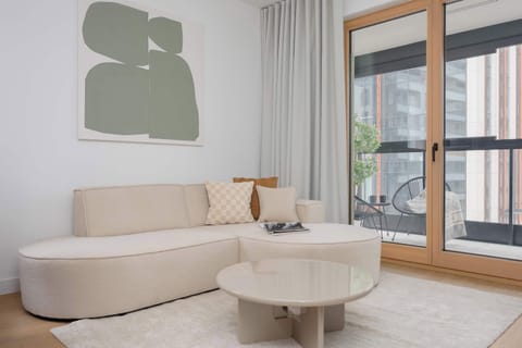 A bright living room with a cream-colored sofa, round coffee table, and large windows. Soft green wall art adds a modern touch and natural light fills the space.
