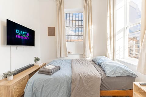 Bright, sunlit bedroom with blue bedding, a wood nightstand, and a TV