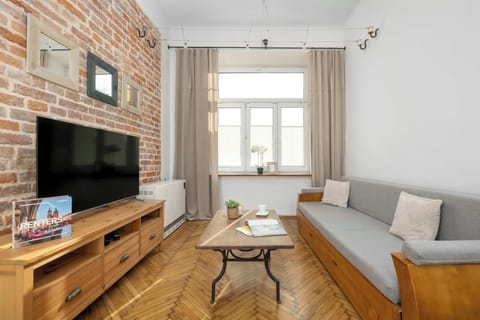 A living room with a grey sofa, wooden furniture, and a TV set. The parquet flooring and brick wall add warmth and texture.