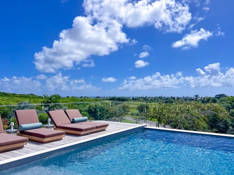 An elegant infinity pool surrounded by plush sun loungers, opening onto a sweeping panoramic view of lush tropical landscape and bright blue sky. A serene outdoor escape designed for ultimate relaxation.