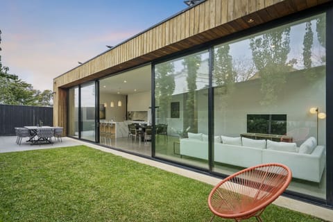 Floor-to-ceiling glass doors open the living and dining space to a private grassed backyard. Relax in the lounge chair or gather at the outdoor dining table as the timber-clad home glows at dusk.