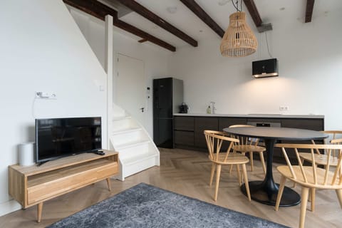Cozy open-plan living area featuring a TV, modern wooden dining set, and fully equipped kitchen — all under warm wooden ceiling beams with natural light pouring in.