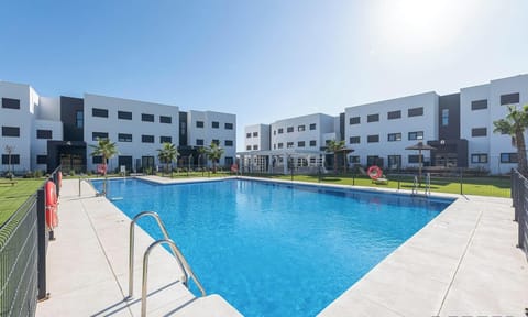 A large, bright blue communal swimming pool is set in a spacious sun deck surrounded by manicured lawns. Modern, multi-story apartment buildings with clean lines frame this attractive, resort-style amenity.