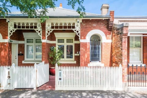 FRONT: Charming red brick Victorian house with white trim, stained glass windows, and a white picket fence. Elegant fretwork and street greenery add to this home's cozy, well-kept appeal.