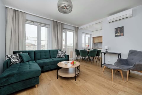 A bright living room with a green sofa, gray armchairs, and a circular coffee table. Large windows bring in natural light, highlighting the warm wooden floor. Wall décor and a ceiling lamp complete the cozy atmosphere.