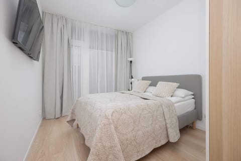 A cozy bedroom featuring a double bed with neutral bedding and soft lighting. The room has light-colored floors and sheer curtains that allow natural light to filter in.