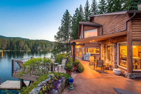 Twilight log-cabin exterior with waterfront patio, gas grill, dock access, and glowing mountain views