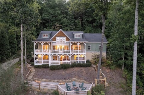 A stunning multi-level mountain home surrounded by tall pines. Warm interior lighting glows through expansive windows, and a large outdoor firepit area sits below, showcasing the property’s serene wooded setting.